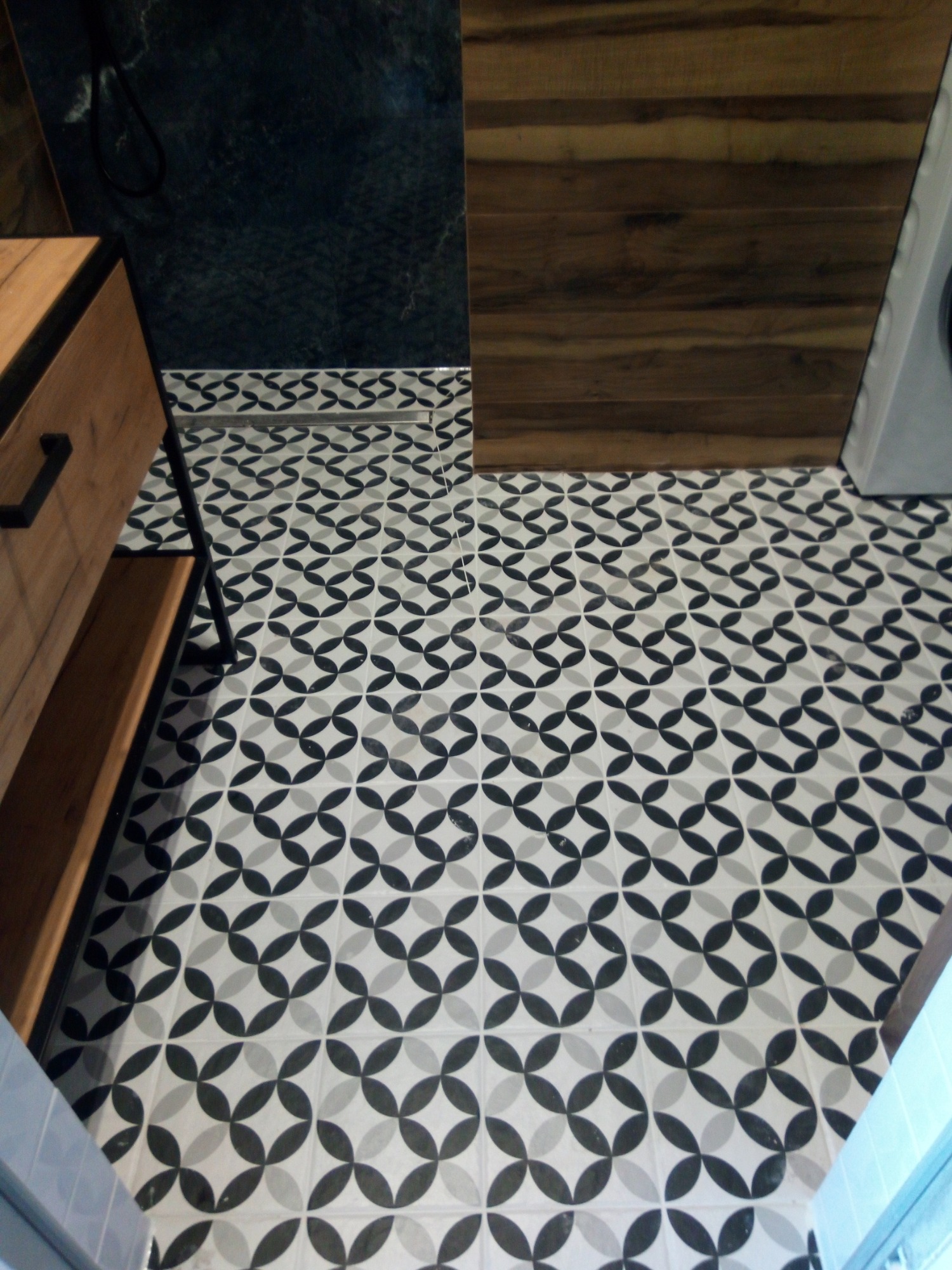 Wzorzysta podłoga łazienki z czarno-białymi płytkami w geometryczny deseń, widoczny fragment drewnianej szafki z czarnymi uchwytami i ciemna ściana w tle.