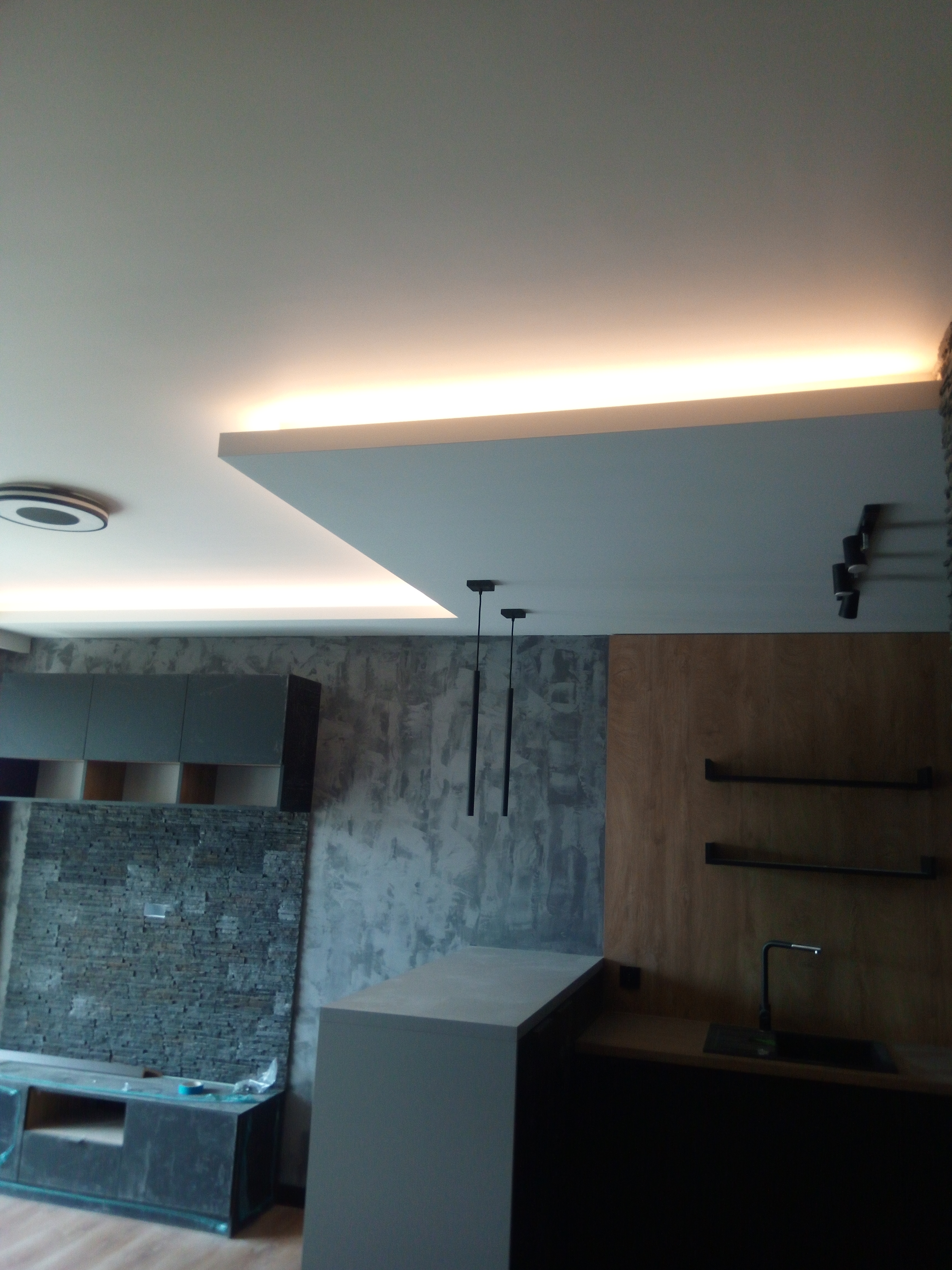 Nowoczesna kuchnia z oświetleniem LED podsufitowym, dekoracyjną ścianą z kamienia i surowym wykończeniem ściany, minimalistyczne czarne lampy wiszące, drewniana zabudowa z półkami.