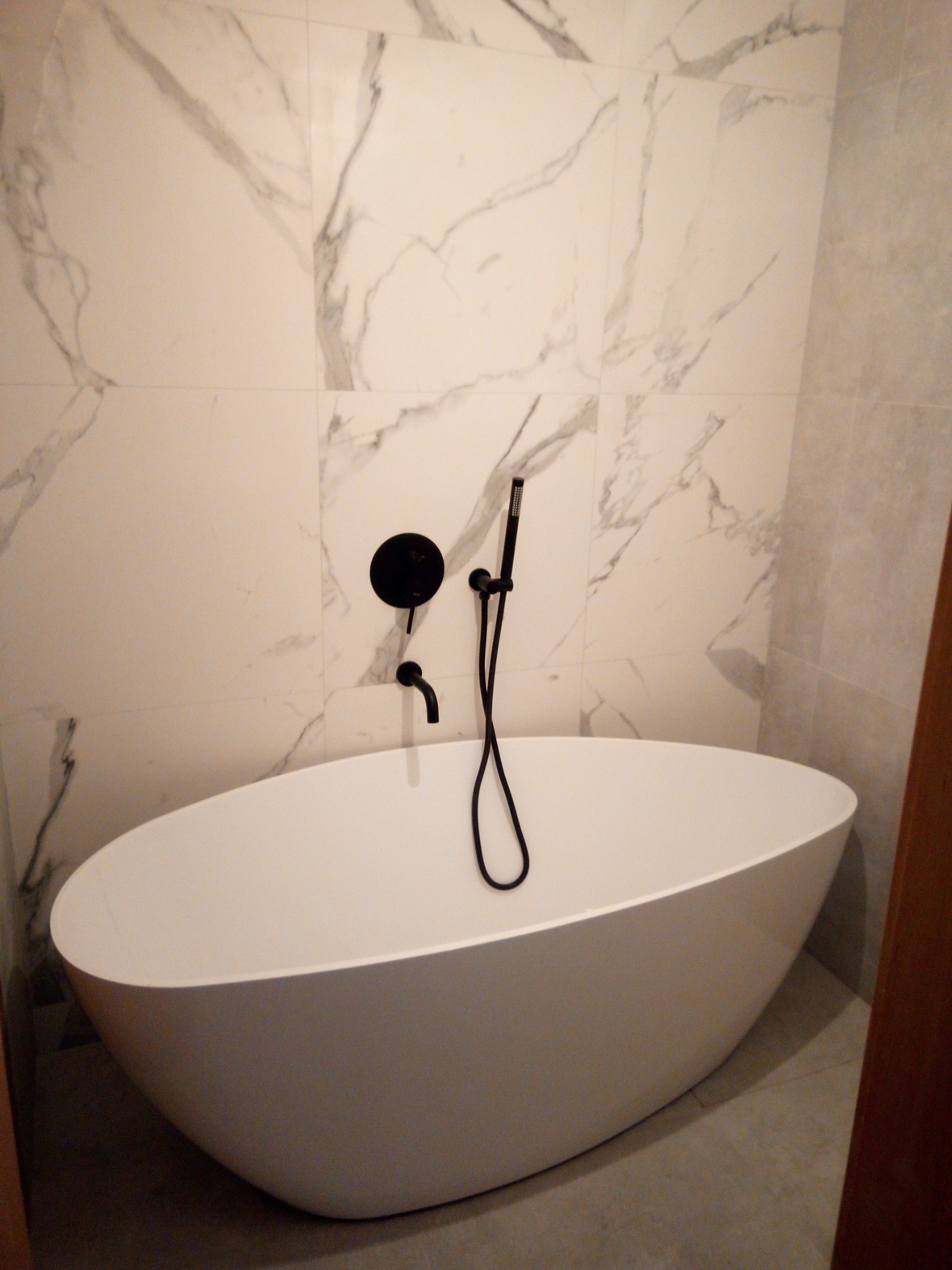 Wolnostojąca wanna o owalnym kształcie z czarną armaturą na tle ściany wyłożonej płytkami imitującymi marmur.