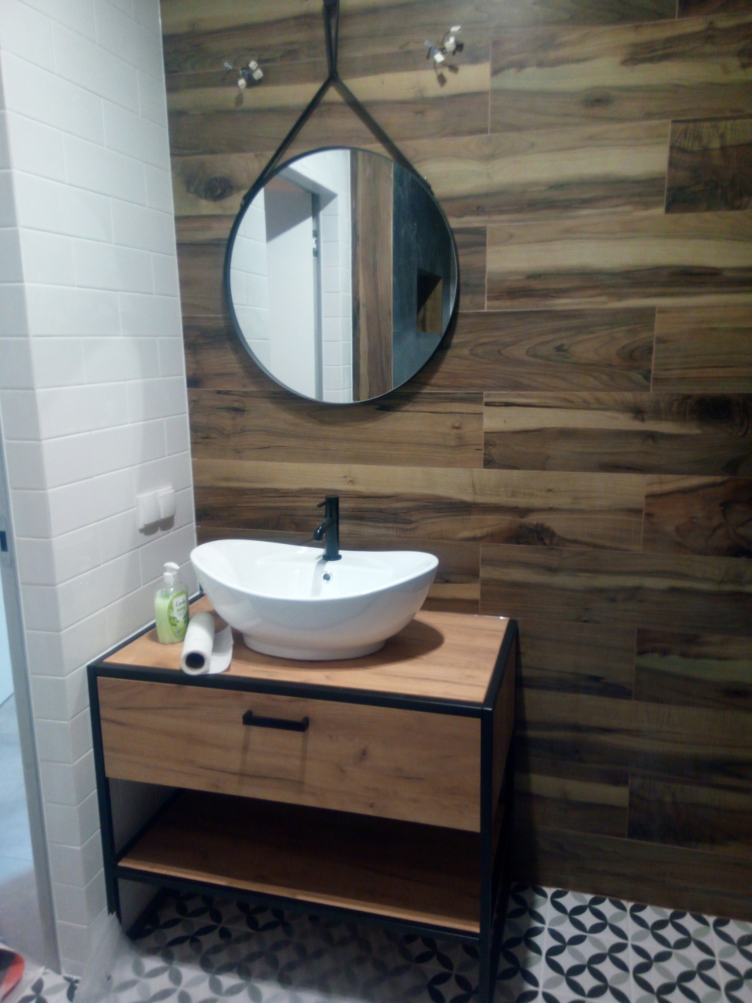 Nowoczesna łazienka z okrągłym lustrem na tle drewnopodobnych płytek, biała umywalka nablatowa na drewnianej szafce z czarnymi elementami, wzorzysta podłoga.