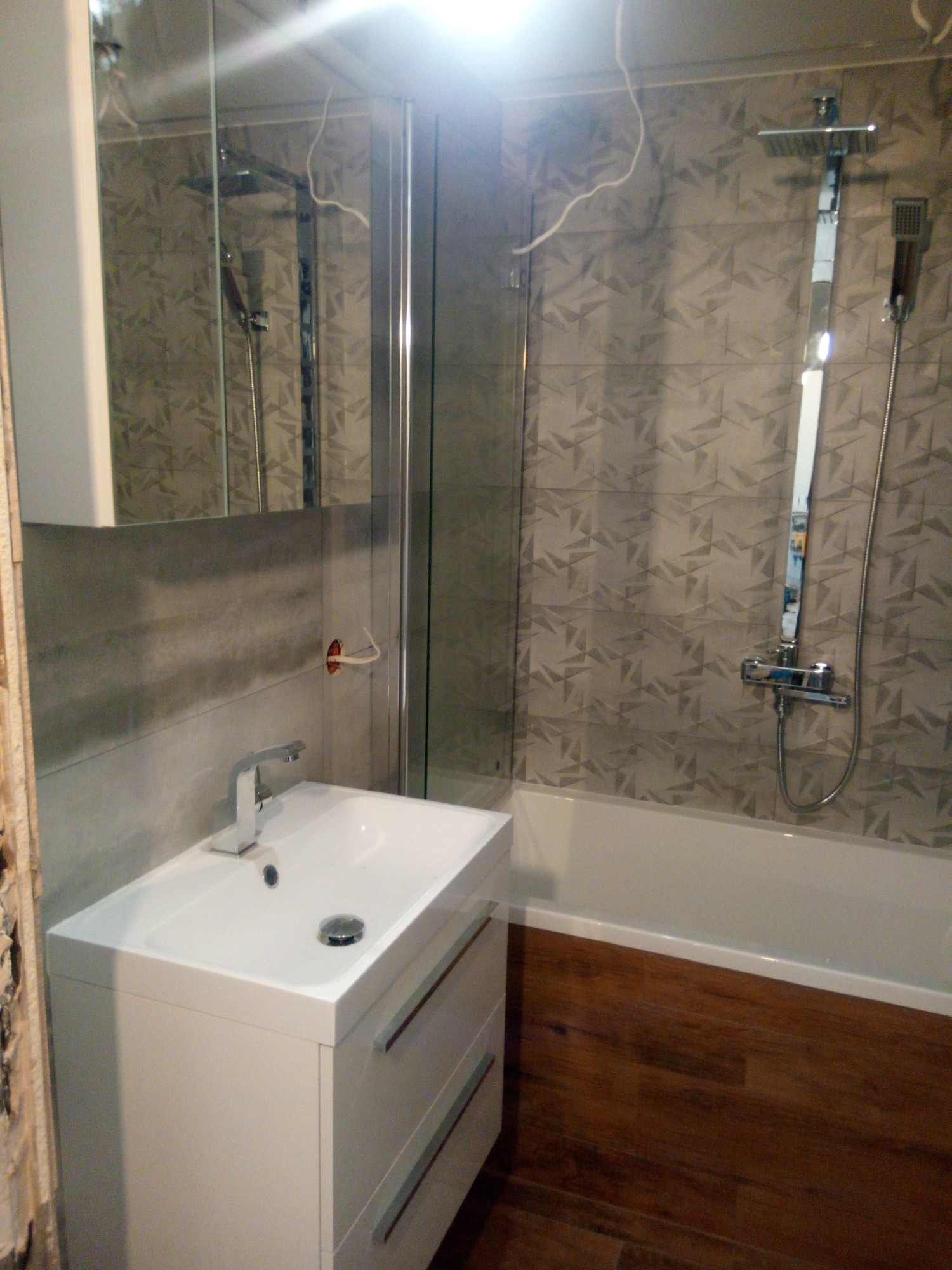 Nowoczesna łazienka z umywalką na szafce z szufladami, kabiną prysznicową i panelami imitującymi drewno na wannie, widoczna instalacja elektryczna przed wykończeniem.