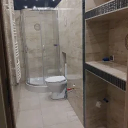 Wykończona łazienka z kabiną prysznicową, toaletą i półkami wyłożonymi mozaiką, płytki w odcieniach beżu z marmurkowym wzorem, widoczne przyłącza hydrauliczne.