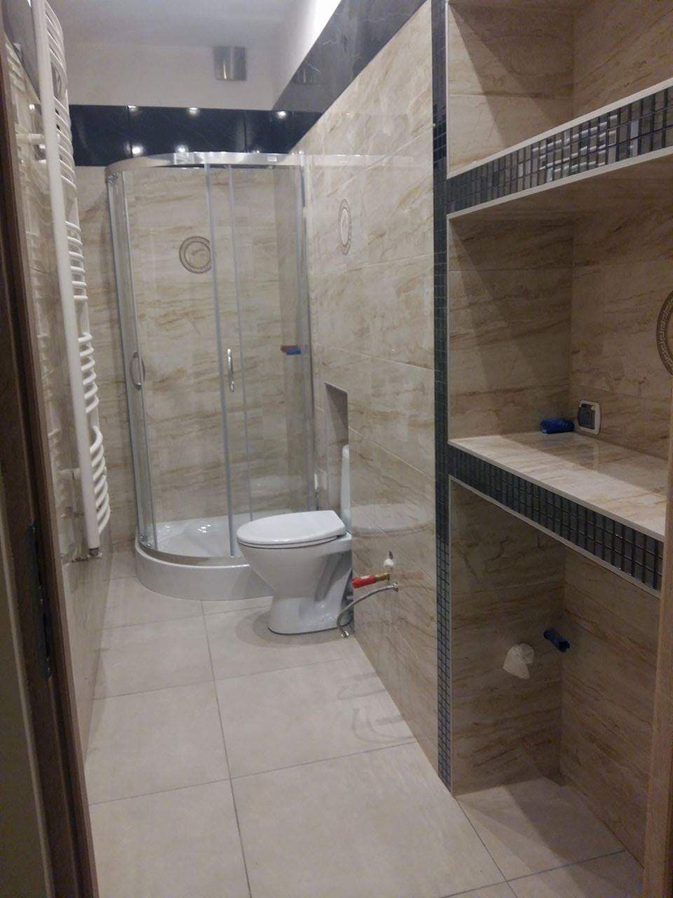 Wykończona łazienka z kabiną prysznicową, toaletą i półkami wyłożonymi mozaiką, płytki w odcieniach beżu z marmurkowym wzorem, widoczne przyłącza hydrauliczne.