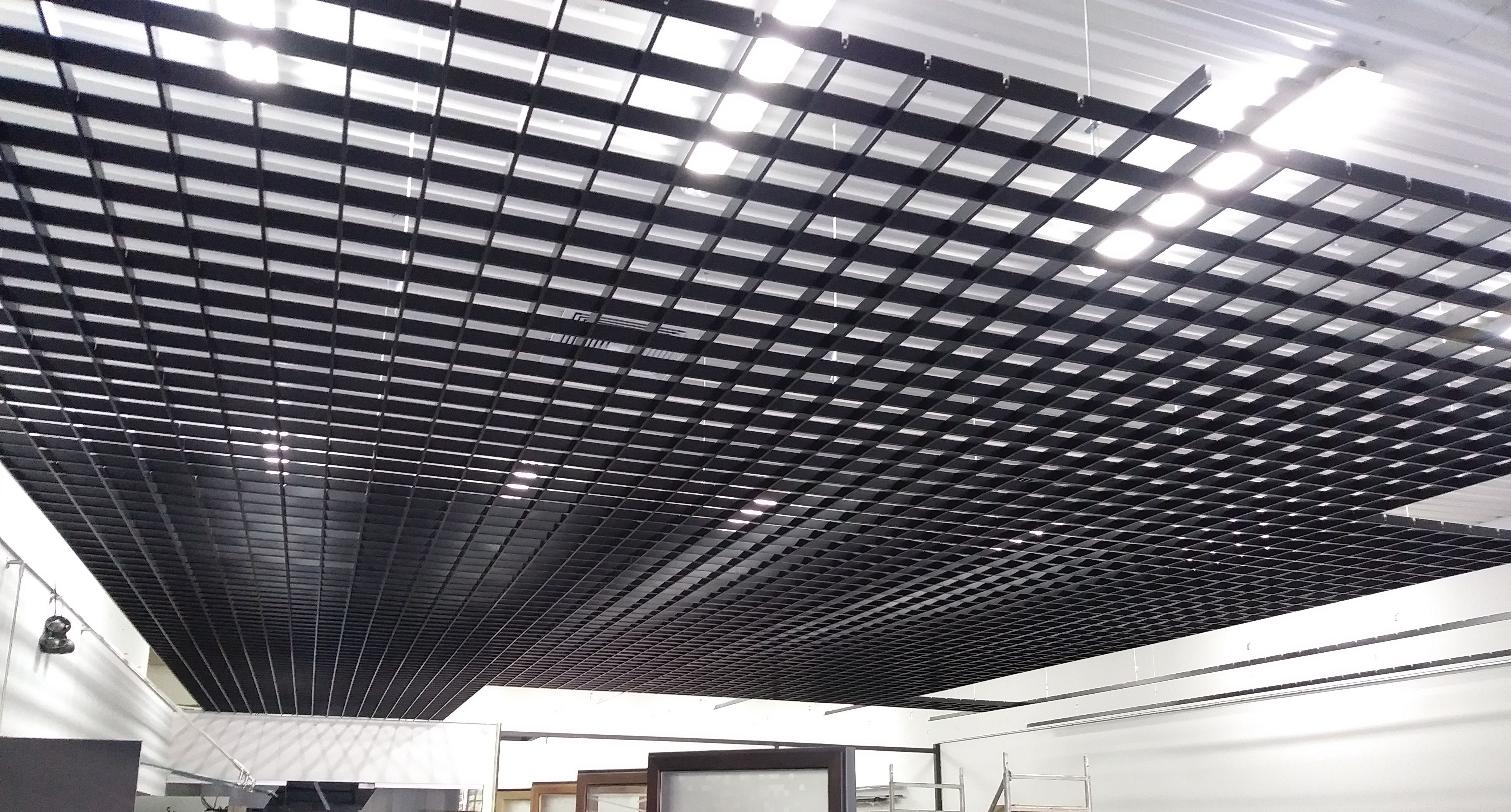 Czarna, kratowa konstrukcja sufitu podwieszanego z widocznymi lampami rastrowymi, oświetlającymi wnętrze. Elementy konstrukcyjne zawieszone na linkach.