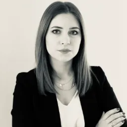 Portret kobiety w czarnym żakiecie i białej bluzce, z rękami skrzyżowanymi na piersiach, na jasnym tle. Widoczny zegarek na nadgarstku i delikatna biżuteria.