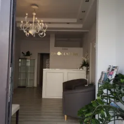 Jasne wnętrze recepcji z widocznym biurkiem, krzesłami, dekoracyjnym żyrandolem i roślinami, widziane z zewnątrz przez otwarte drzwi.