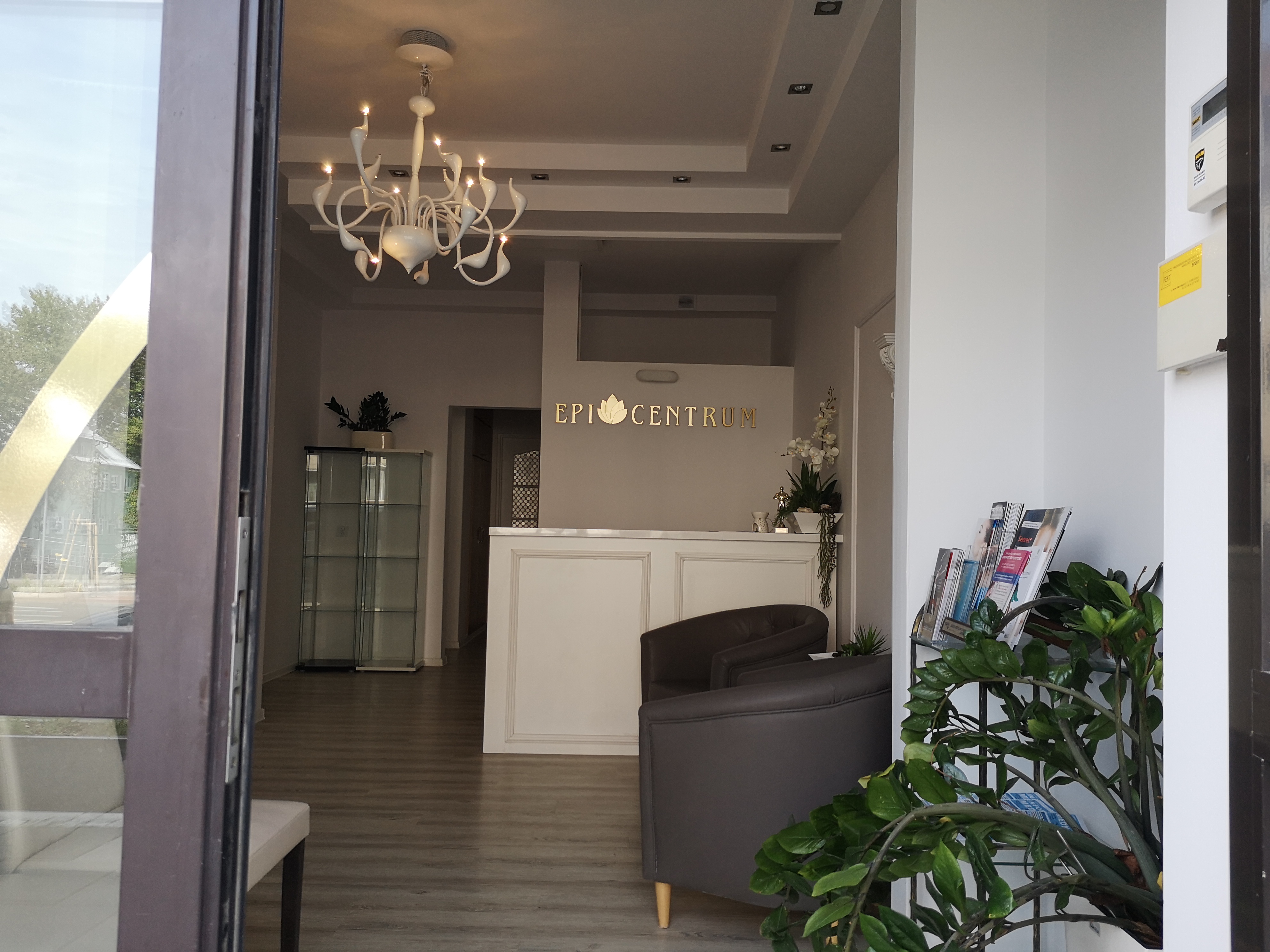 Jasne wnętrze recepcji z widocznym biurkiem, krzesłami, dekoracyjnym żyrandolem i roślinami, widziane z zewnątrz przez otwarte drzwi.