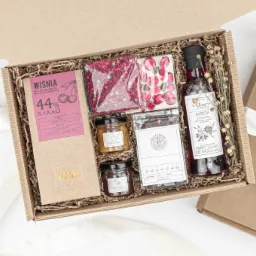 Otwarty kartonowy prezentowy box wypełniony czekoladą wiśniową, syropem z kwiatów, konfiturą pomarańczową i truskawkową z migdałami, oraz czekoladą z malinami, obok zamknięte pudełko z kokardą.