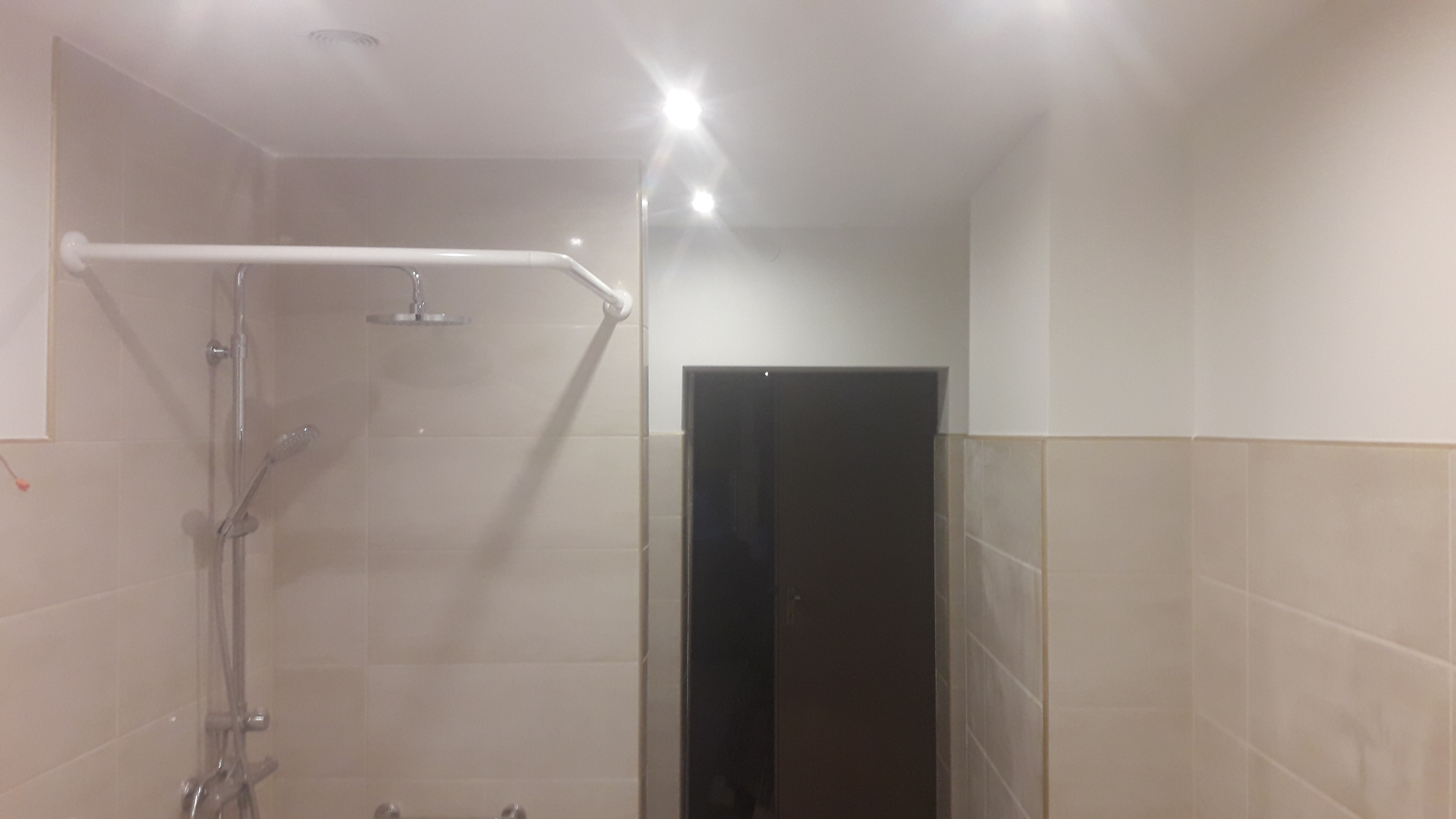 Wykończona łazienka z beżowymi płytkami, kabiną prysznicową z deszczownicą i uchwytem dla niepełnosprawnych, oświetlona punktowymi lampami sufitowymi.