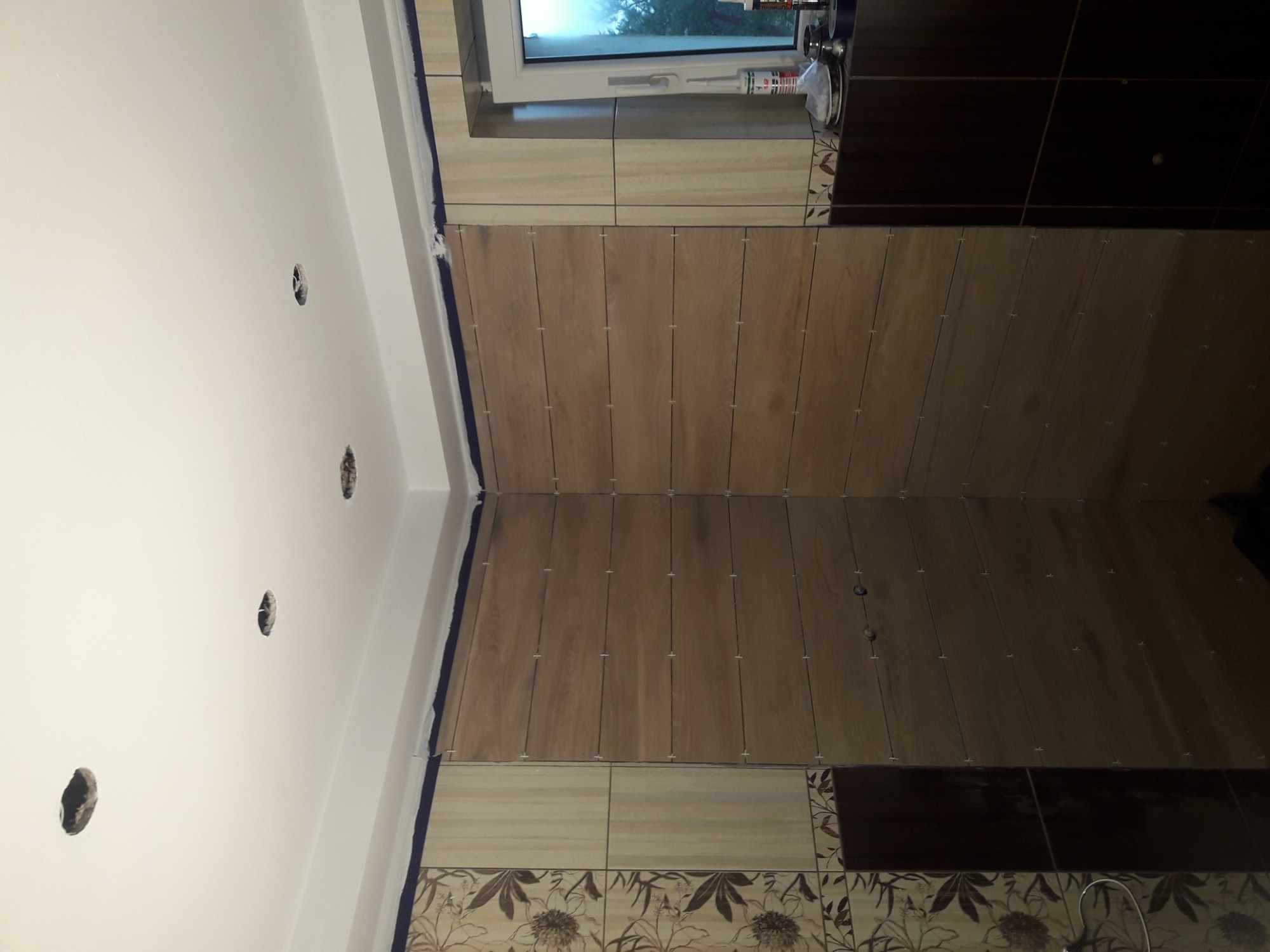 Fragment ściany w trakcie układania płytek imitujących drewno, widoczne krzyżyki dystansowe, górna część ściany pomalowana na biało z wyciętymi otworami na gniazdka, zabezpieczona taśmą malarską.