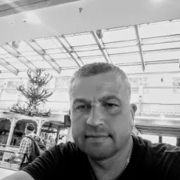 Czarno-białe selfie mężczyzny z widocznym żyrandolem z poroża i fragmentem przeszklonego dachu w tle, sugerujące wnętrze dworca lub lotniska.