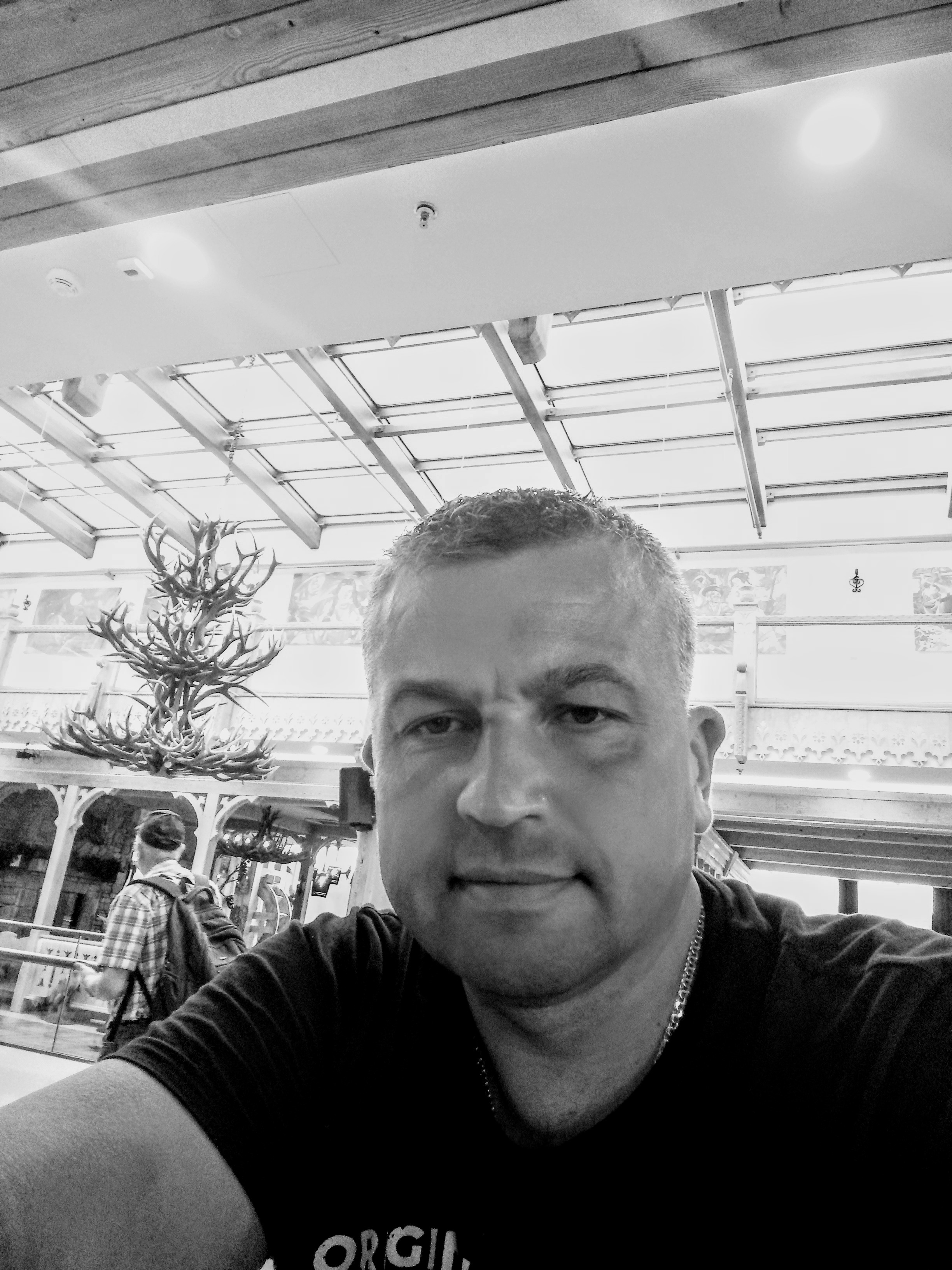 Czarno-białe selfie mężczyzny z widocznym żyrandolem z poroża i fragmentem przeszklonego dachu w tle, sugerujące wnętrze dworca lub lotniska.