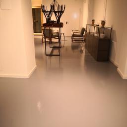 SPIC AND SPAN. Home & Office Cleaning - Minimalistyczne wnętrze biurowe z szarą, połyskliwą podłogą, nowoczesnymi meblami i oświetleniem, krzesła ustawione na stole, sugerujące porządkowanie przestrzeni.