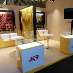 SPIC AND SPAN. Home & Office Cleaning - Stoisko targowe z żółtymi akcentami: podświetlane gabloty z logo firmy, plakaty z produktami spożywczymi i bydłem, białe krzesła odwrócone na stołach, czarny telewizor na ścianie.