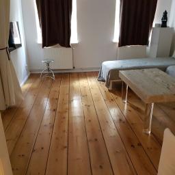 SPIC AND SPAN. Home & Office Cleaning - Jasny pokój dzienny z drewnianą podłogą, minimalistycznym stolikiem kawowym z przezroczystymi nogami, szarymi sofami i brązowymi zasłonami przy oknach.