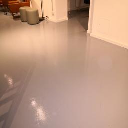 SPIC AND SPAN. Home & Office Cleaning - Błyszcząca, szara posadzka w jasnym wnętrzu biurowym z widocznymi meblami wypoczynkowymi i minimalistycznymi pufami w tle.