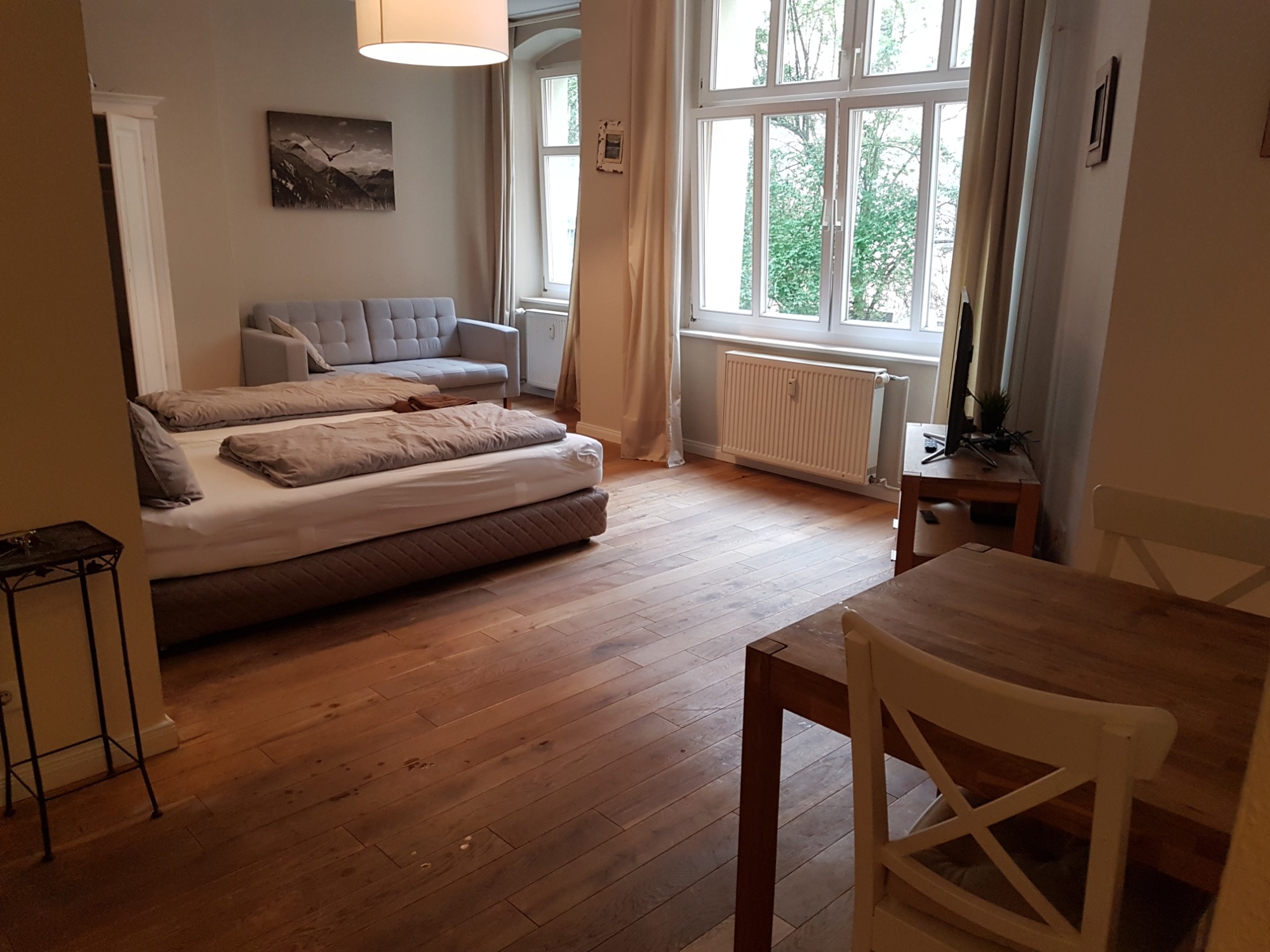 Jasne wnętrze pokoju z dużym oknem, drewnianą podłogą i łóżkiem z beżową pościelą, widoczne także stół z krzesłami i szara sofa.