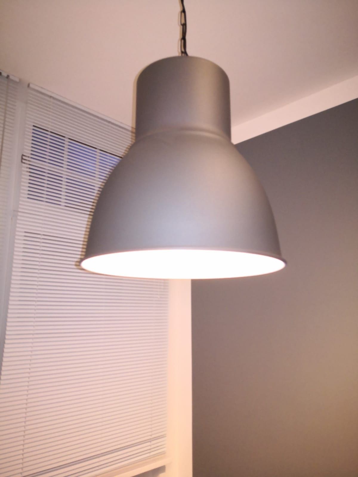 Szara lampa wisząca z łańcuchem, oświetlająca pomieszczenie z żaluzjami i szarą ścianą.