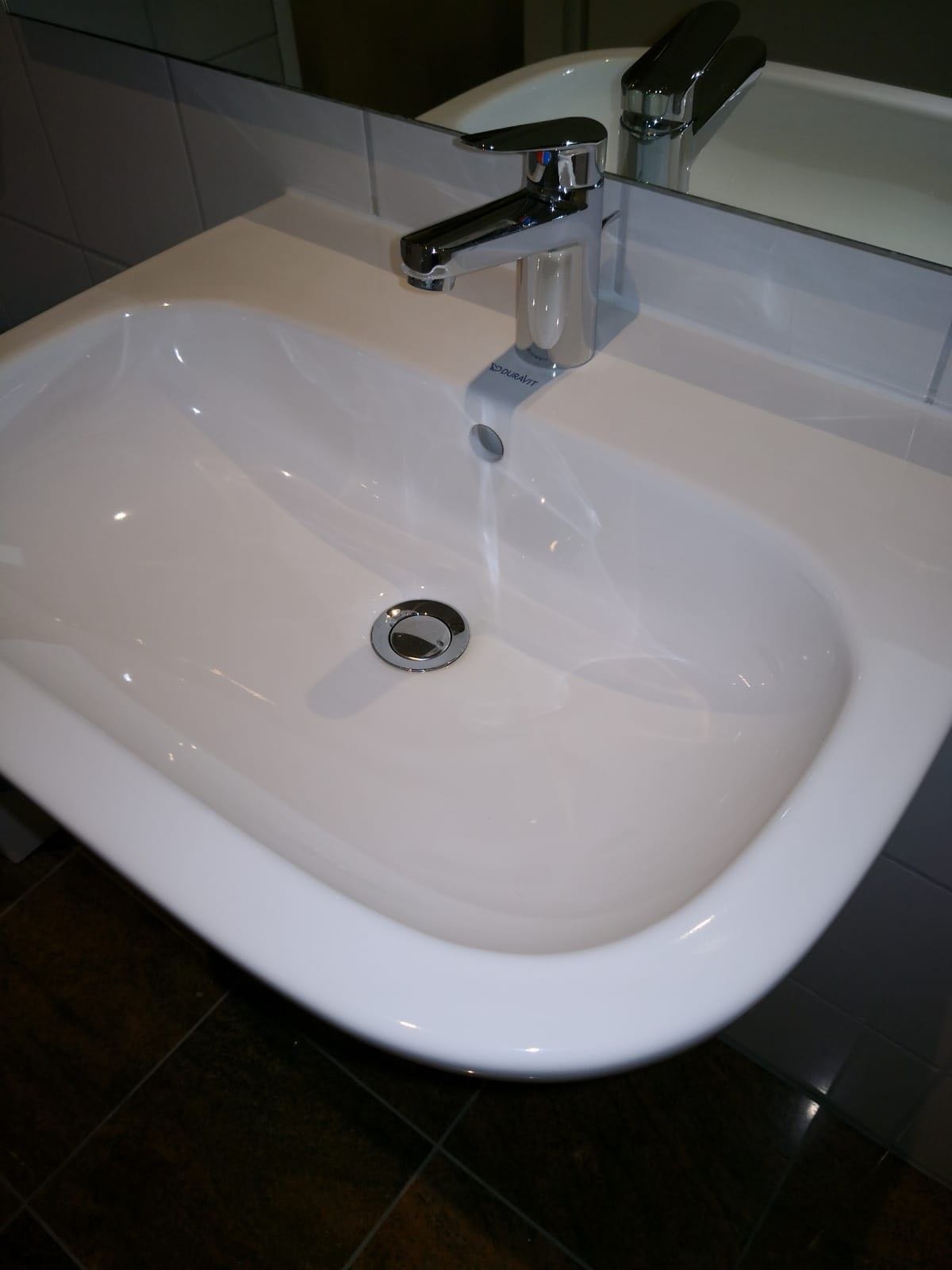 Biała umywalka Duravit z chromowaną baterią i odpływem, odbijająca się w lustrze na tle płytek.