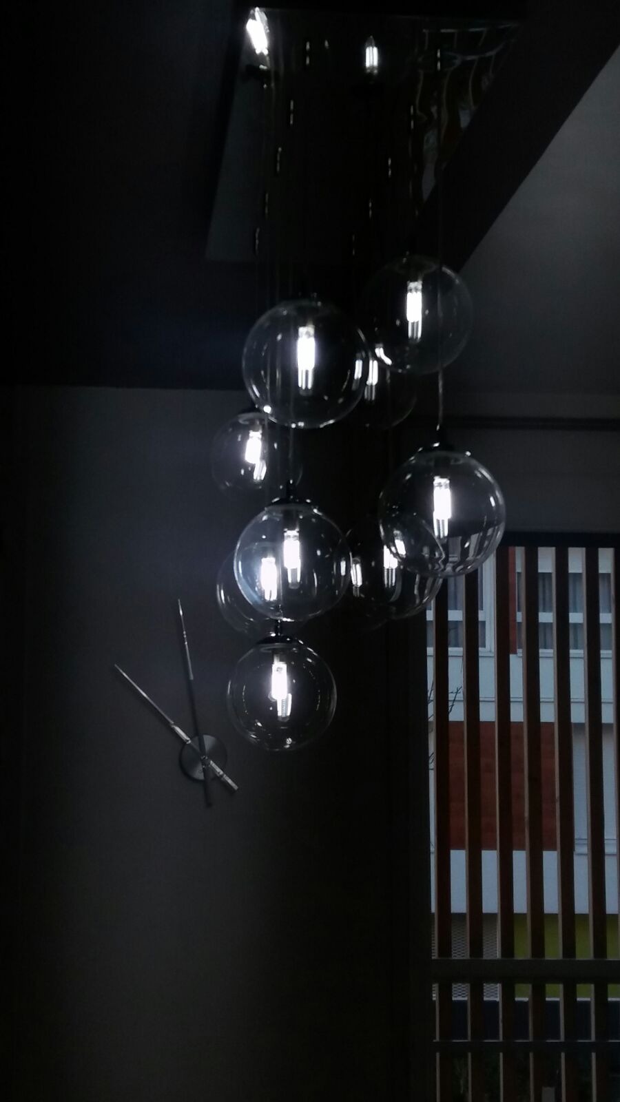 Szklana lampa sufitowa w kształcie kuli z żarówkami LED, wisząca nad ścianą z nowoczesnym zegarem i oknem z drewnianymi żaluzjami w tle.