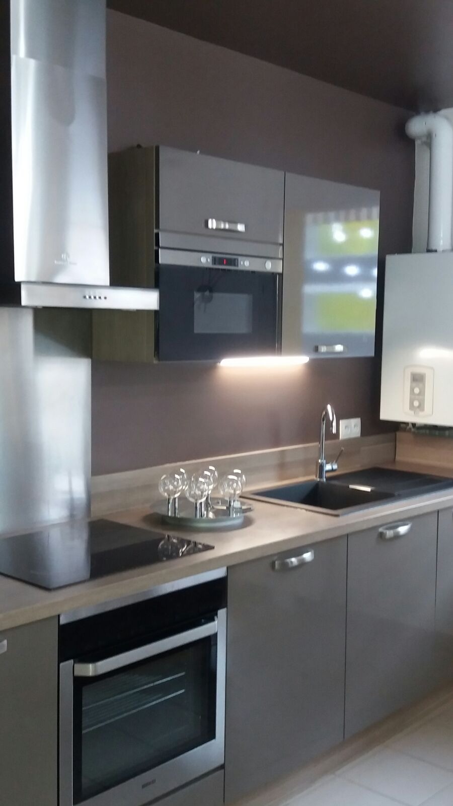 Nowoczesna kuchnia w odcieniach szarości i brązu z zabudową kuchenną, płytą indukcyjną, piekarnikiem, zlewem i baterią, oświetleniem podszafkowym oraz dekoracyjnymi szklanymi kulami na blacie.