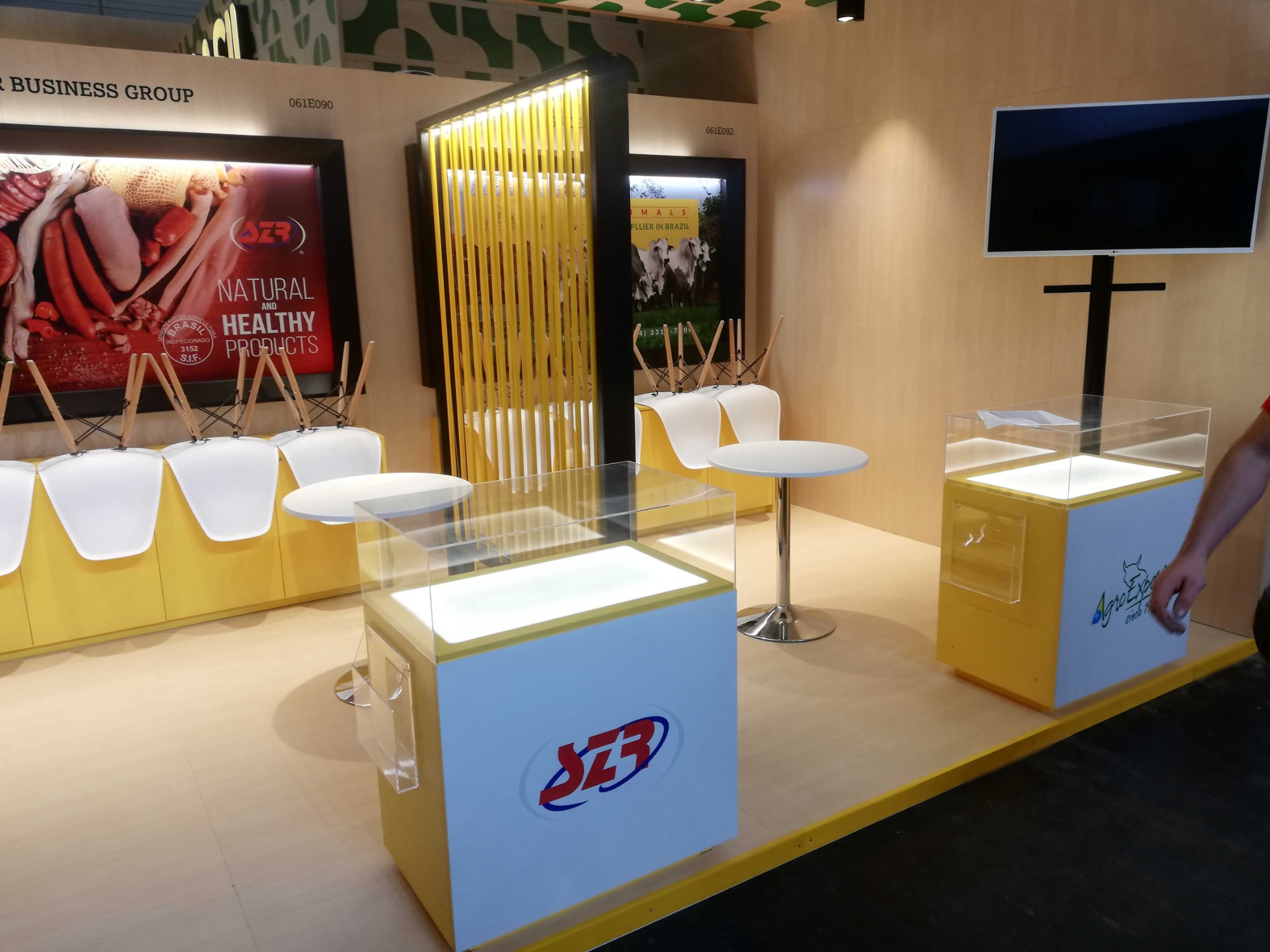 Stoisko targowe z żółtymi akcentami: podświetlane gabloty z logo firmy, plakaty z produktami spożywczymi i bydłem, białe krzesła odwrócone na stołach, czarny telewizor na ścianie.