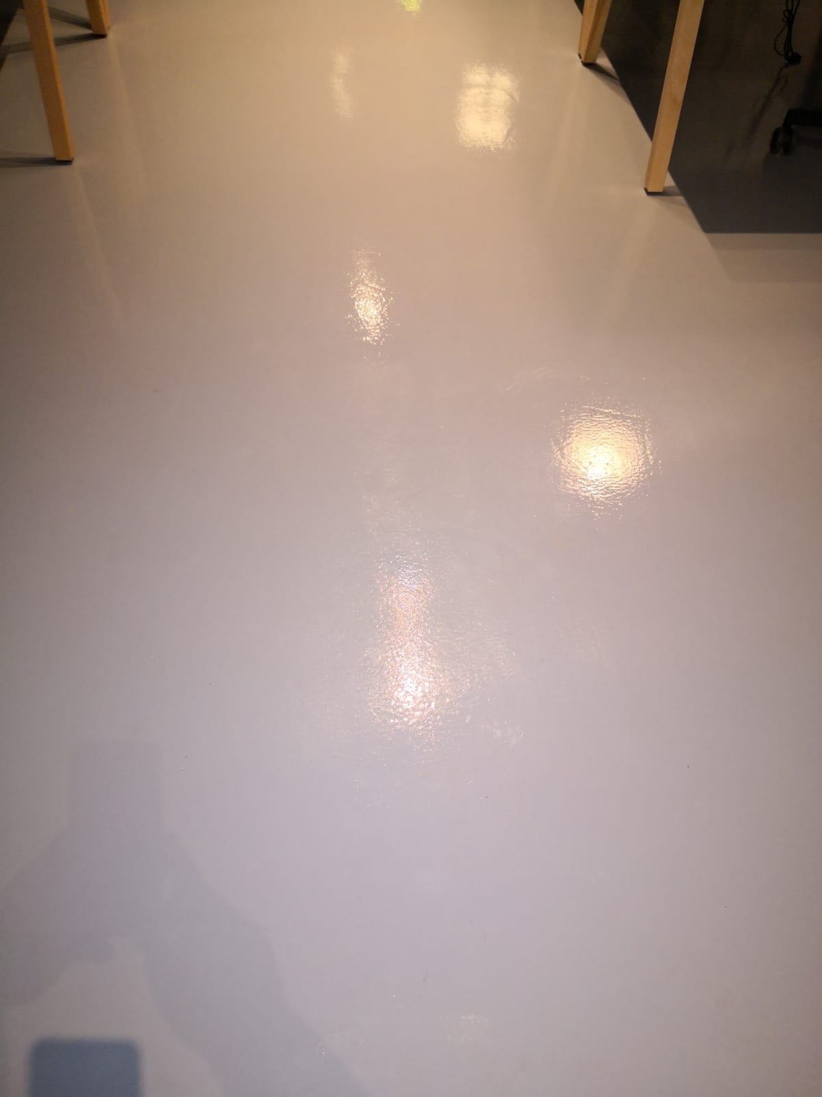 Błyszcząca, jasnoszara podłoga po umyciu, z widocznymi refleksami światła i fragmentami nóg biurek.