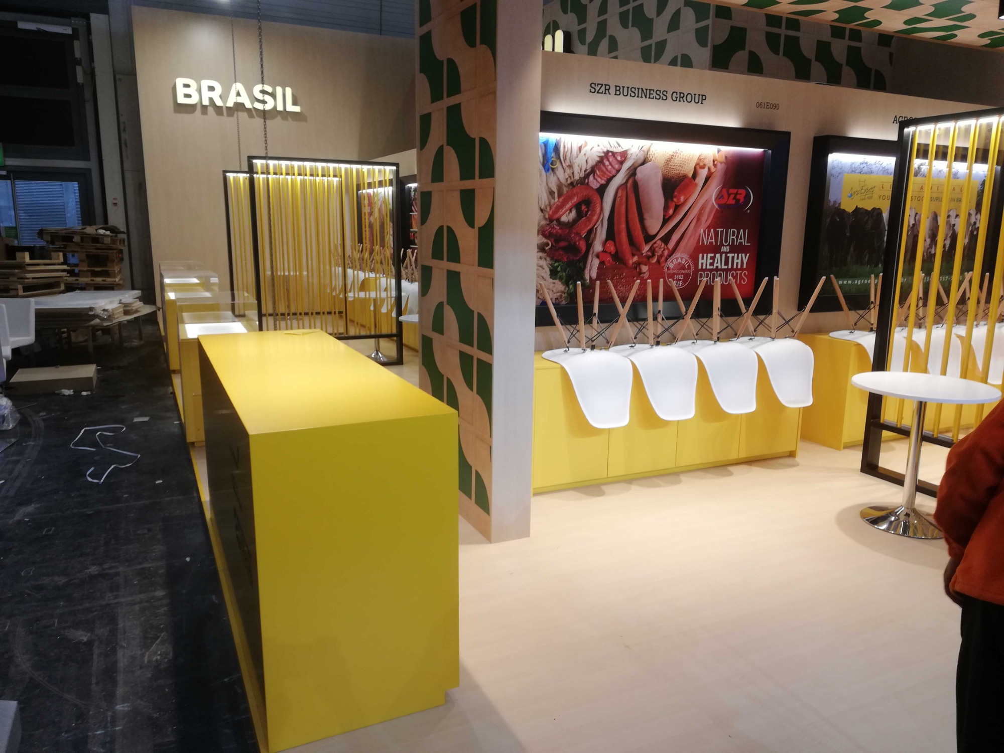 Żółte stoisko targowe z napisem BRASIL i logo SZR Business Group, ekspozycja produktów spożywczych na plakacie, krzesła oparte o ladę, przygotowania do targów, widoczne palety.