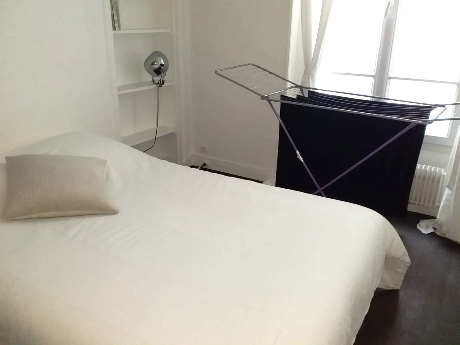 Jasna sypialnia z białym łóżkiem, srebrną lampą na ścianie i suszarką na pranie z czarnymi ubraniami przy oknie.