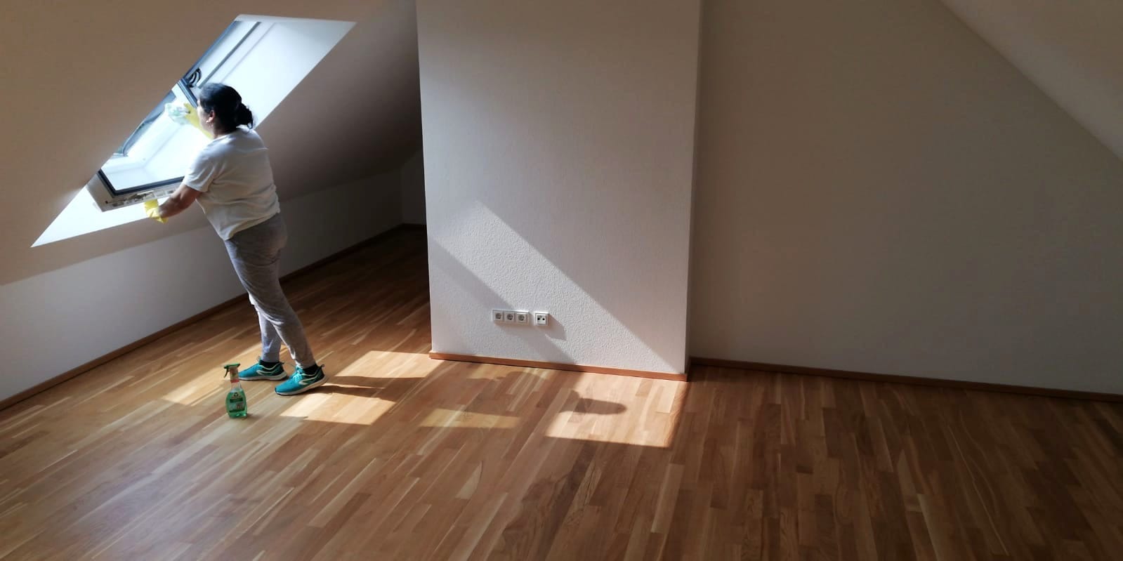 Osoba w szarych spodniach i białej koszulce myje okno dachowe w pustym pokoju; na podłodze stoi butelka ze środkiem czyszczącym.