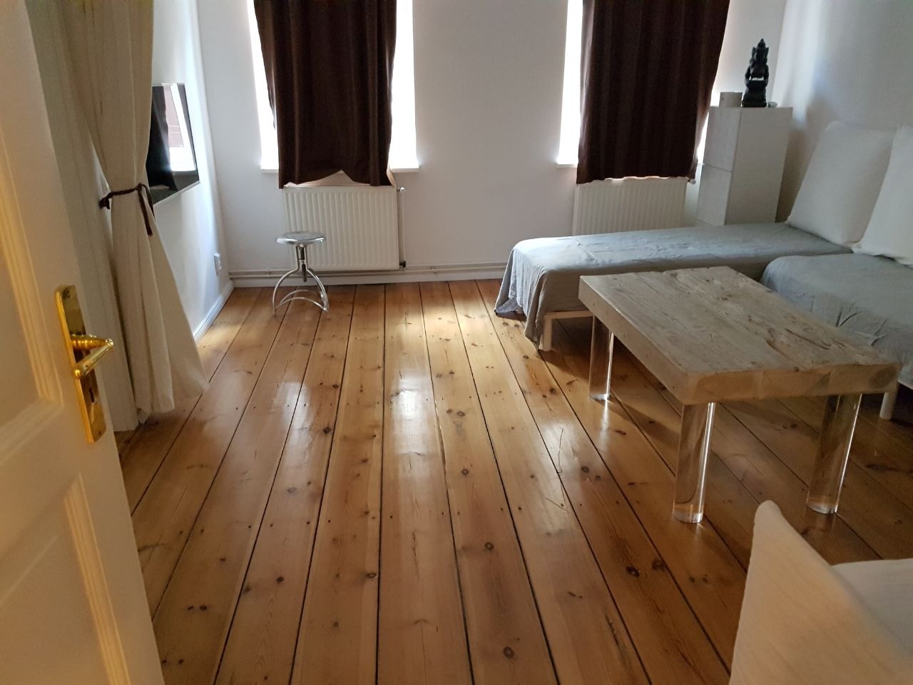 Jasny pokój dzienny z drewnianą podłogą, minimalistycznym stolikiem kawowym z przezroczystymi nogami, szarymi sofami i brązowymi zasłonami przy oknach.