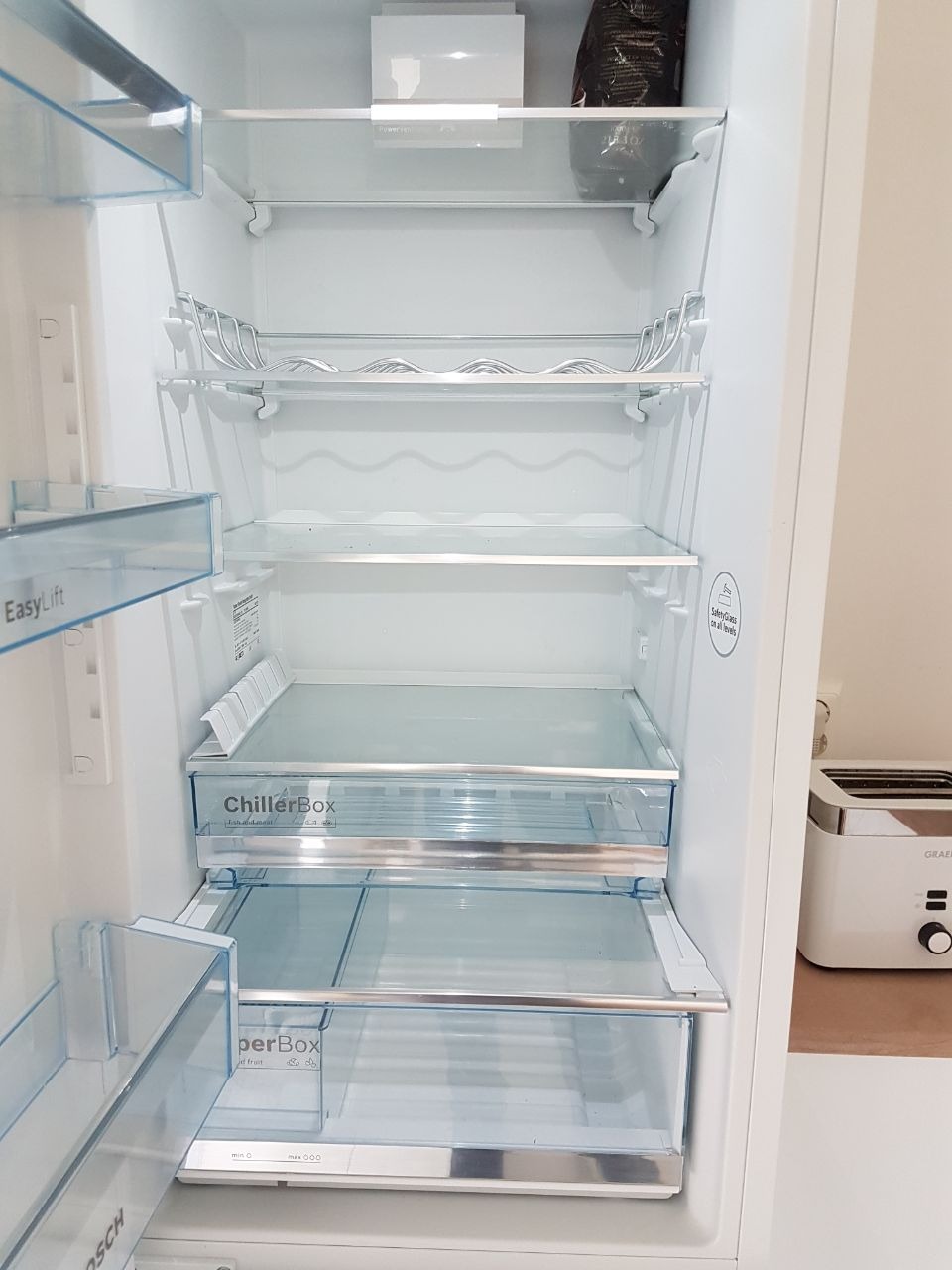 Pusta, czysta lodówka marki Bosch z półkami, szufladami ChillerBox i SuperBox, obok toster na blacie.