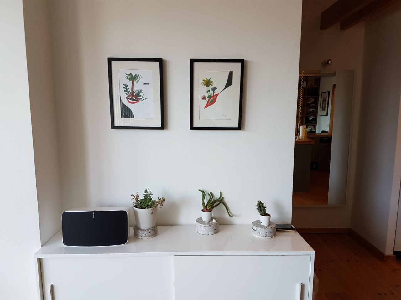 Biała komoda z dwoma obrazkami w czarnych ramkach, trzema małymi doniczkami z sukulentami na betonowych podstawkach i głośnikiem Sonos. W tle widoczne wejście do garderoby z lustrem.