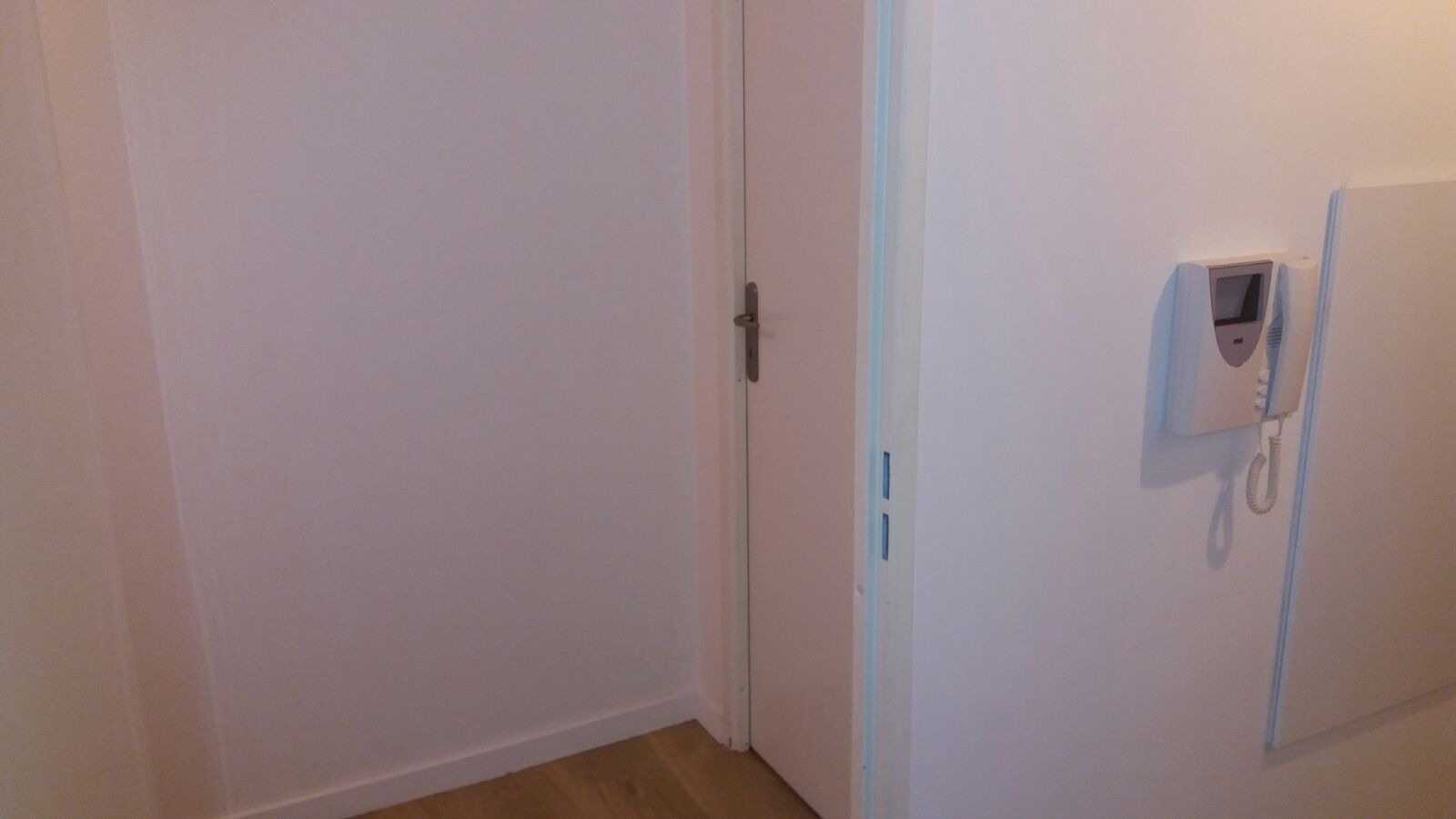 Jasne wnętrze z drzwiami i domofonem na ścianie, widoczne ślady po czyszczeniu na ścianie po lewej stronie.