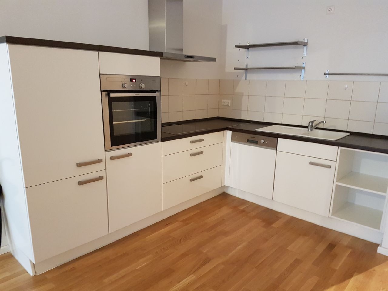 Jasna kuchnia z białymi szafkami i drewnianą podłogą, widoczny piekarnik, zlew i okap, na ścianie płytki i metalowe półki.