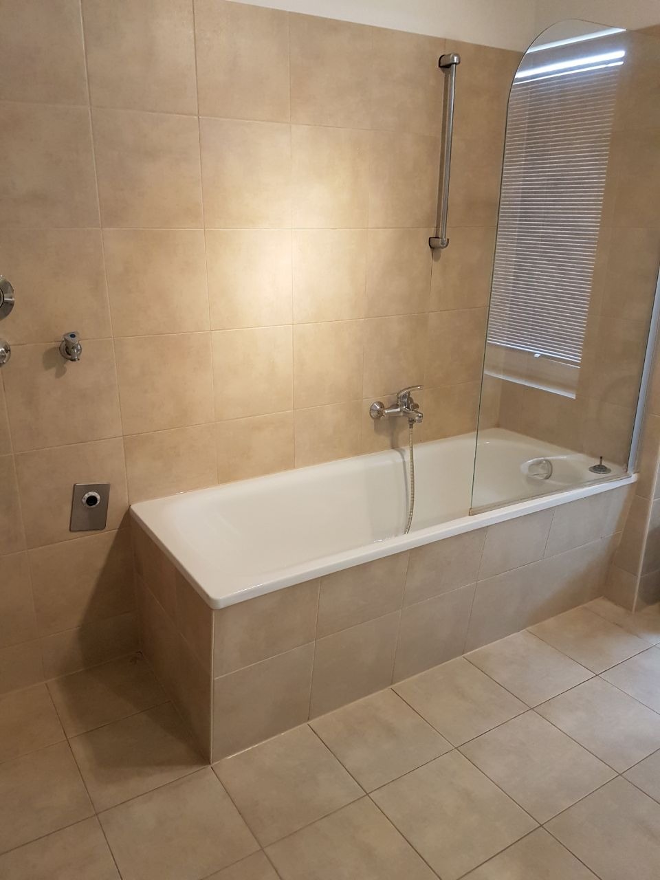 Wanna obudowana beżowymi płytkami, z zamontowaną szklaną osłoną, widok na fragment łazienki z beżowymi płytkami na ścianach i podłodze.