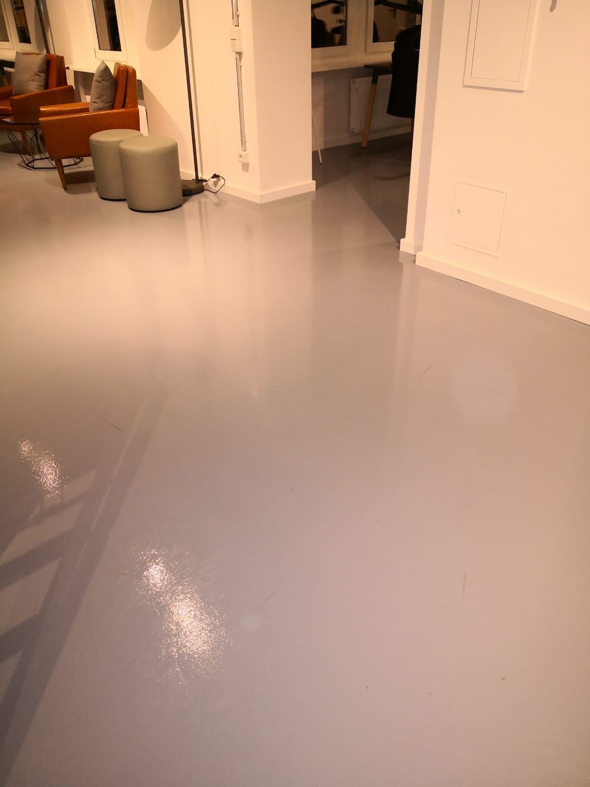 Błyszcząca, szara posadzka w jasnym wnętrzu biurowym z widocznymi meblami wypoczynkowymi i minimalistycznymi pufami w tle.