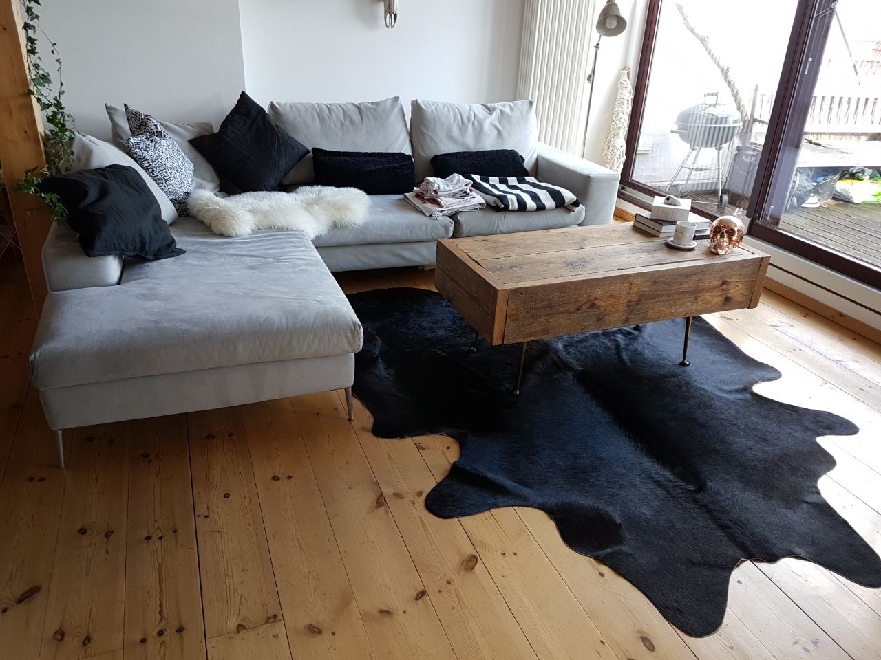 Jasnoszara sofa narożna z czarnymi poduszkami i białym pledem, drewniany stolik kawowy z dekoracyjną czaszką, czarna skóra na podłodze, widok na balkon z grillem.