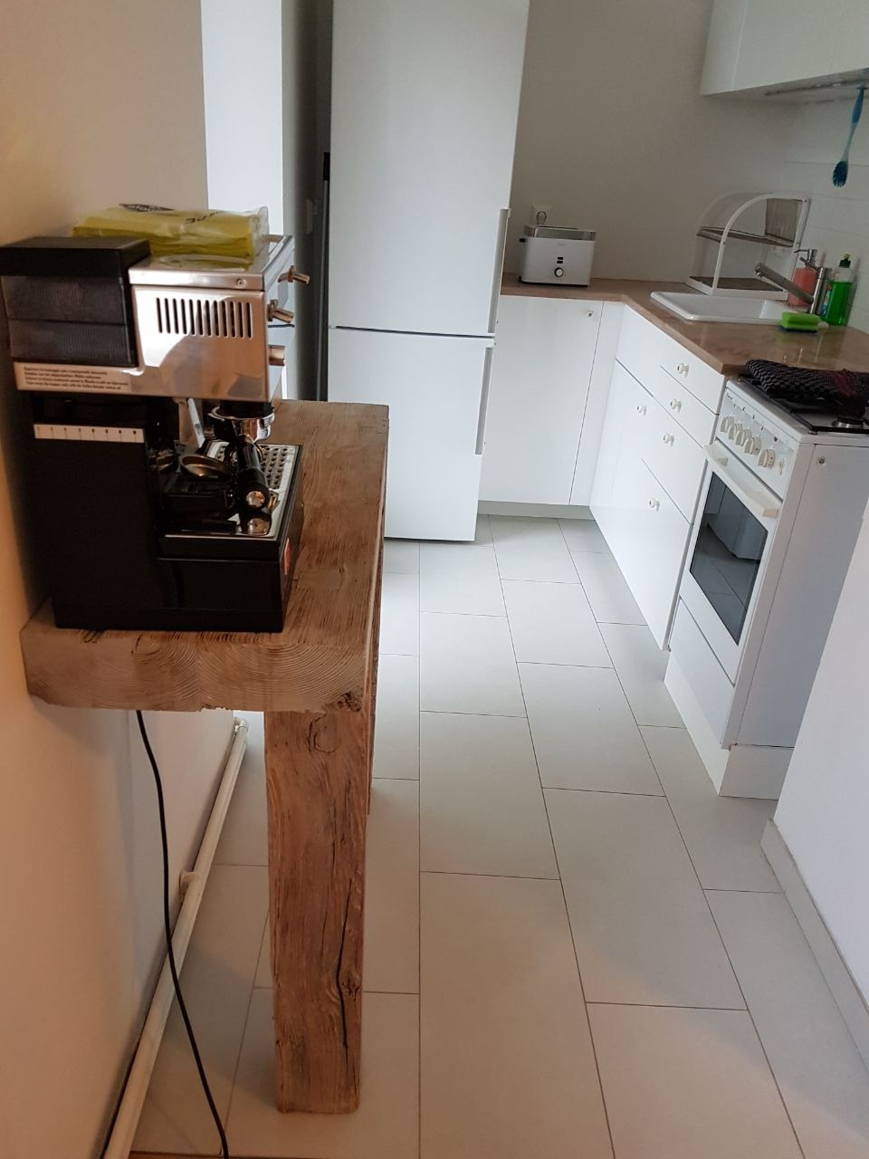 Wnętrze kuchni z minimalistyczną zabudową, białe płytki podłogowe, ekspres do kawy na blacie z surowego drewna.