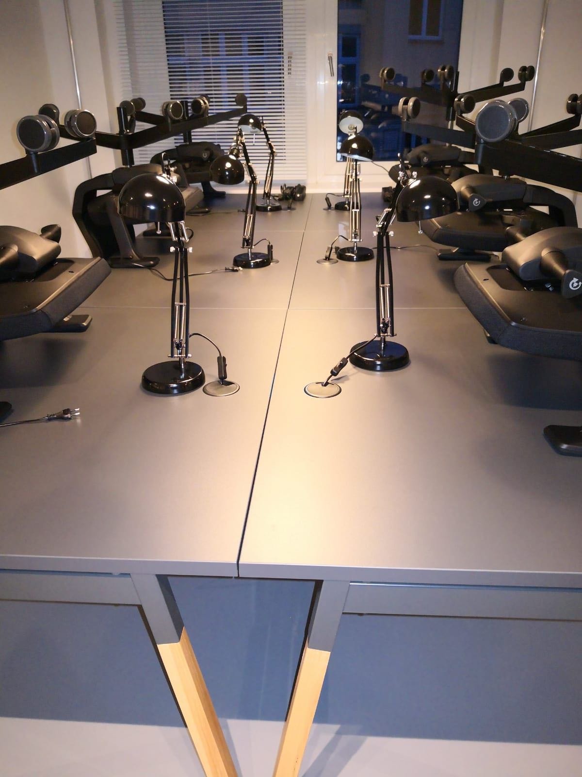 Rząd biurek z szarymi blatami, czarnymi lampkami biurkowymi i ergonomicznymi krzesłami w nowoczesnym biurze, widok z przodu na blaty i lampki.