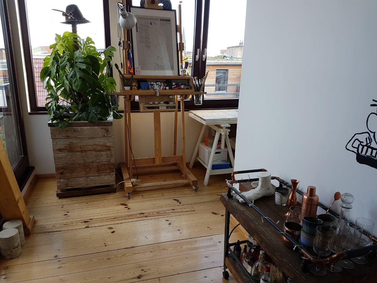 Jasne wnętrze mieszkania z drewnianą podłogą, sztalugą z obrazem, rośliną w drewnianej skrzyni z kapeluszem na szczycie i wózkiem barowym z naczyniami.