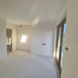 Wnętrze pokoju po pracach przygotowawczych, ściany pokryte białą warstwą, widoczne puszki elektryczne, surowy beton na podłodze, okno z drewnianą ramą wpuszczające naturalne światło.