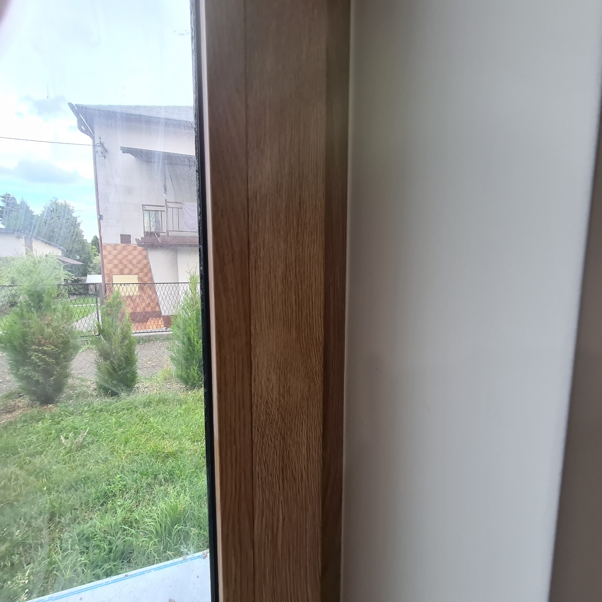 Fragment okna z drewnianą ramą i widokiem na zewnątrz: trawa, krzewy i budynek w tle, ściana pomalowana na biało wewnątrz.