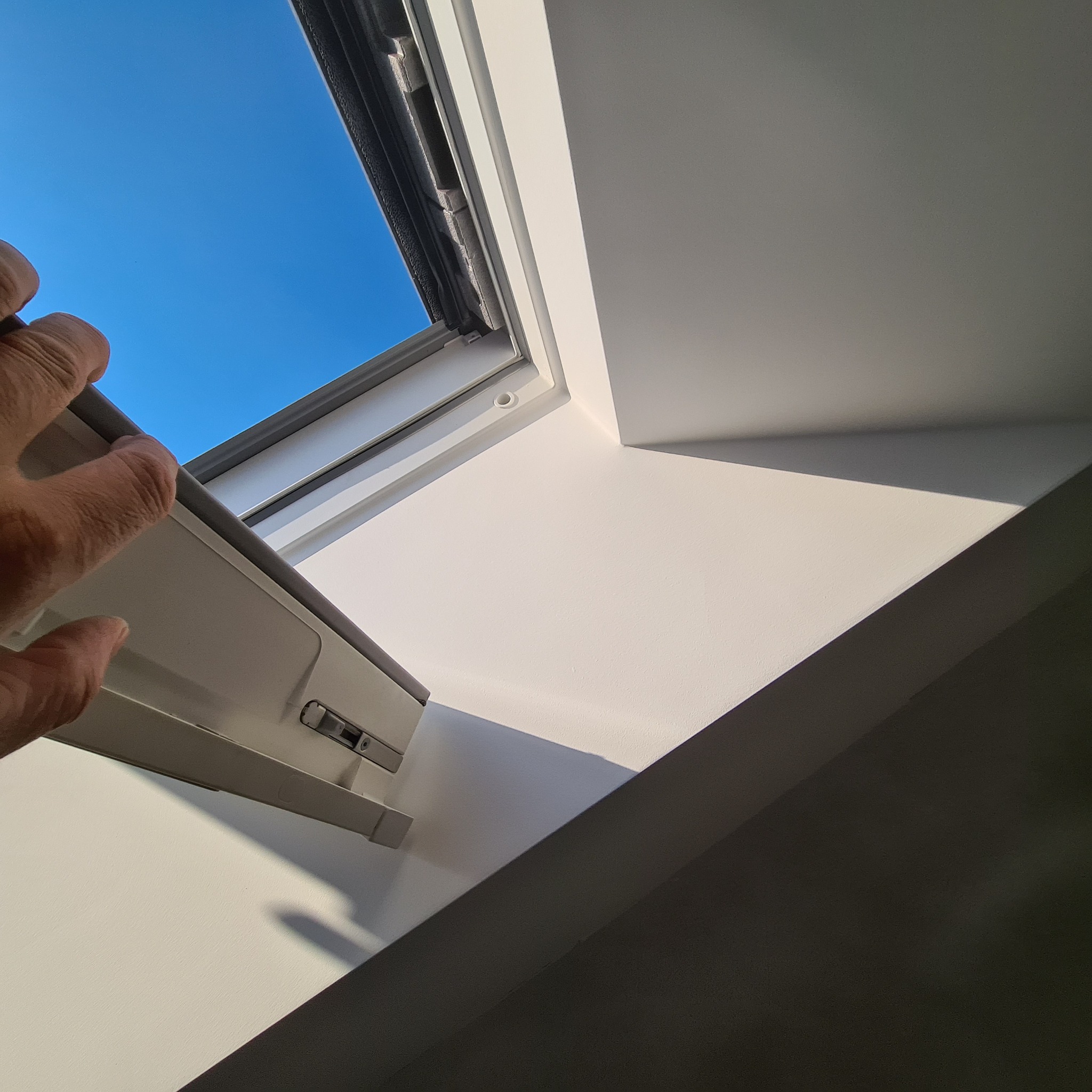 Ręka otwiera okno dachowe, widok na błękitne niebo, gładka, biała powierzchnia ściany i sufitu, kontrastowe cienie.