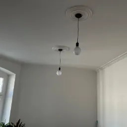 Białe wnętrze po remoncie z dwoma wiszącymi żarówkami na czarnych kablach, dekoracyjnymi rozetami sufitowymi i sztukaterią przy suficie, widok z dołu na sufit i fragment ściany z oknem.