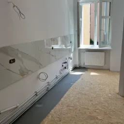 Wnętrze w trakcie prac: ściana z fragmentem marmurowych płytek nad instalacją wodną, odkryte przewody elektryczne, surowa podłoga z płyt OSB, okno z widokiem na budynek.