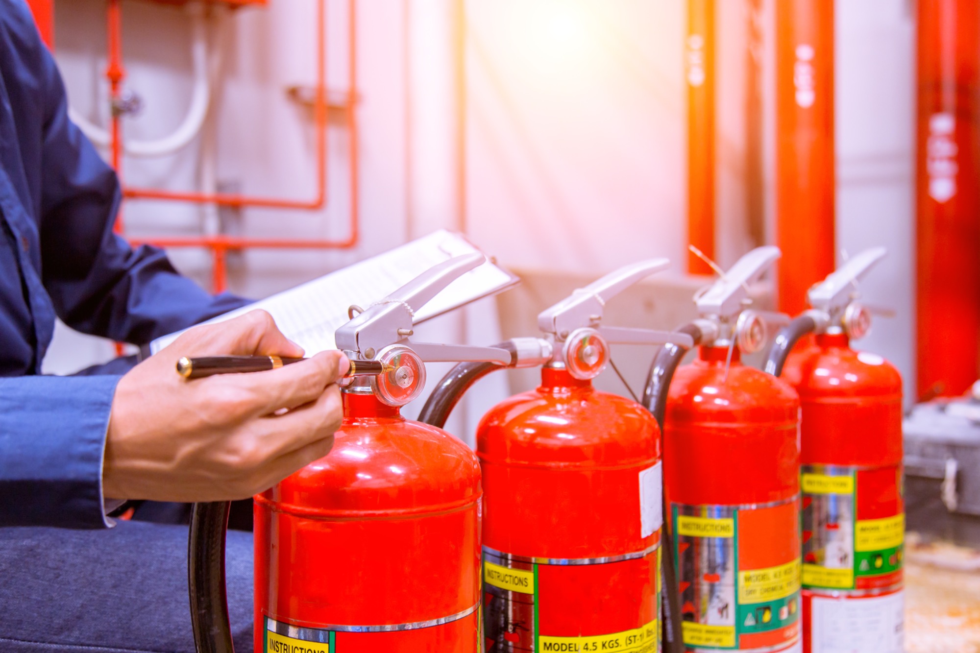 Cztery czerwone gaśnice ustawione w rzędzie, mężczyzna w niebieskiej koszuli sprawdza pierwszą gaśnicę, trzymając w ręku czarny długopis i notatnik.