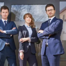Portret zespołu prawnego: dwóch mężczyzn i kobieta w garniturach stoją przed szklanymi drzwiami z napisem 'Radca Prawny' i logo kancelarii.