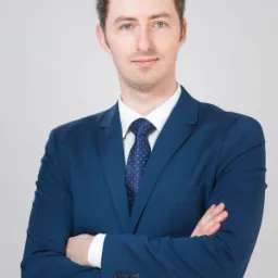 Portret młodego mężczyzny w niebieskim garniturze i kropkowanym krawacie, z założonymi rękami na neutralnym, jasnoszarym tle.