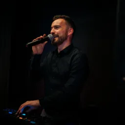 Uśmiechnięty DJ z brodą w czarnej koszuli, trzyma mikrofon i miksuje muzykę na konsoli z niebieskim podświetleniem, w tle ciemna scena.